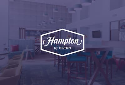 A hotel communal area with the Hampton by Hilton logo layered over the top of the image