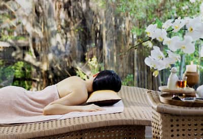 A lady lying on a message table is covered by a towel with her back to the camera. There are massage oils and flowers in the foreground and a tranquil garden in the background. 